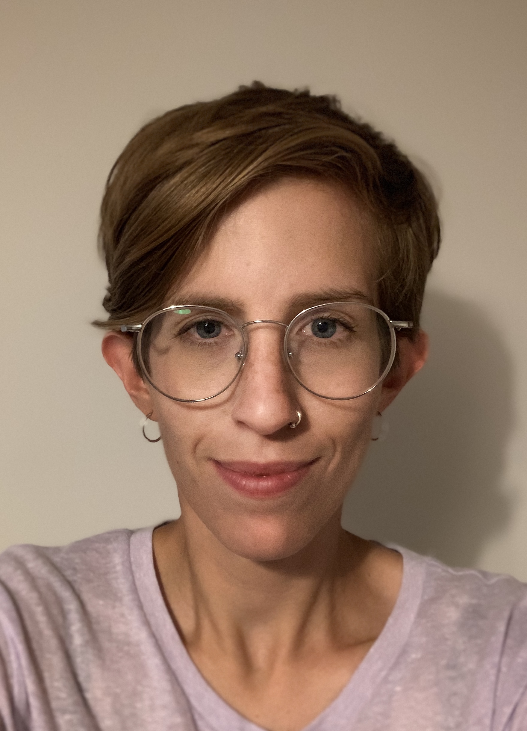 Kat is wearing glasses and a light purple t-shirt. She has short brown hair.