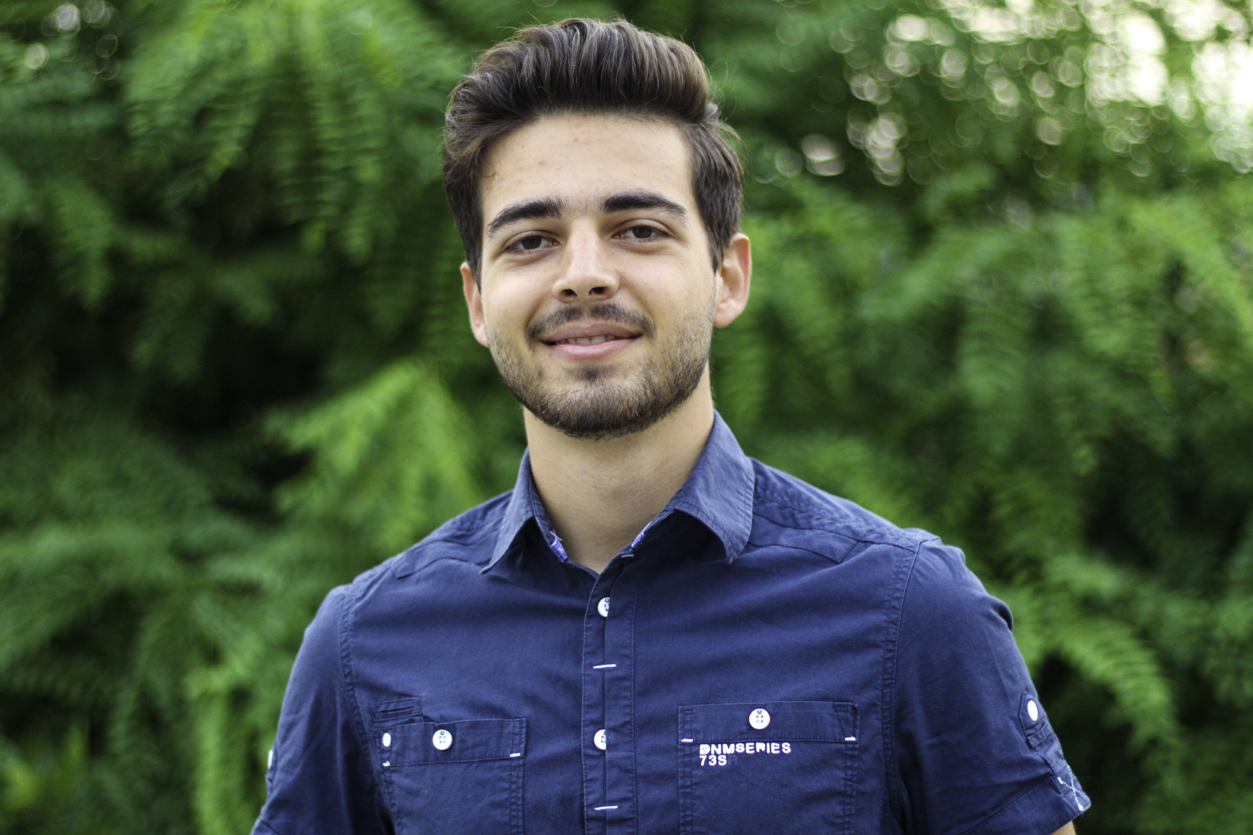 Slavi is wearing a navy button up shirt and is standing in front of a tree.