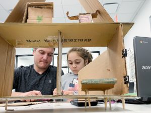 2 people working on a wooden craft while sitting at a desk.