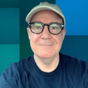 Sean is a white man, wearing dark rimed glasses and a baseball hat. He is looking straight on at the camera.