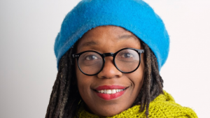 Jheni is looking straight at the camera. She is a black woman, wearing a bright blue hat and a yellow scar. She has round black glasses and red lipstick.